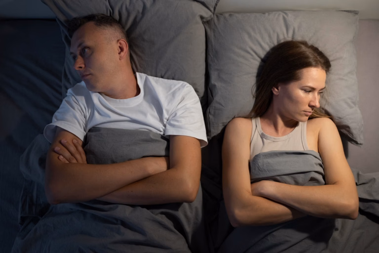 An overhead view of a man and a woman lying in bed on gray pillows and sheets, separated by space and facing away from each other with arms crossed and serious, distant expressions.