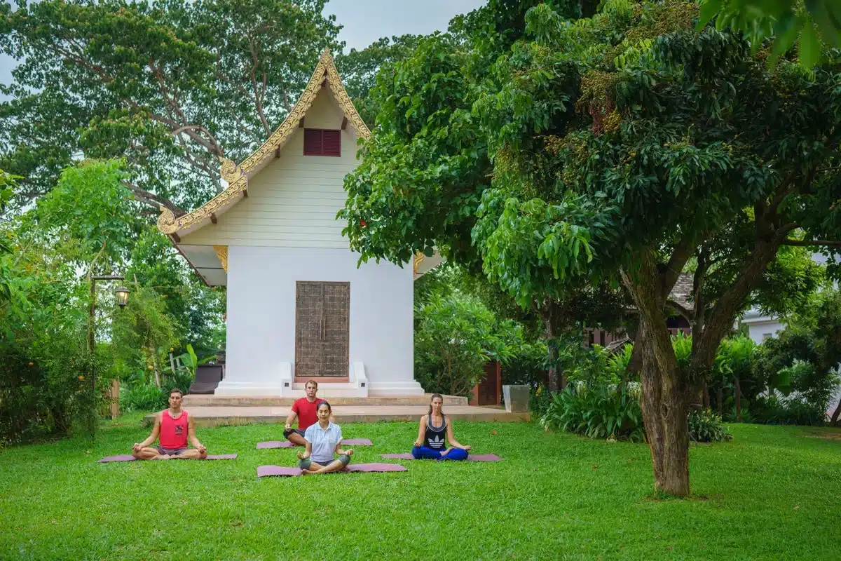 A group of four people meditating on yoga mats in front of a traditional building surrounded by greenery at The Dawn.