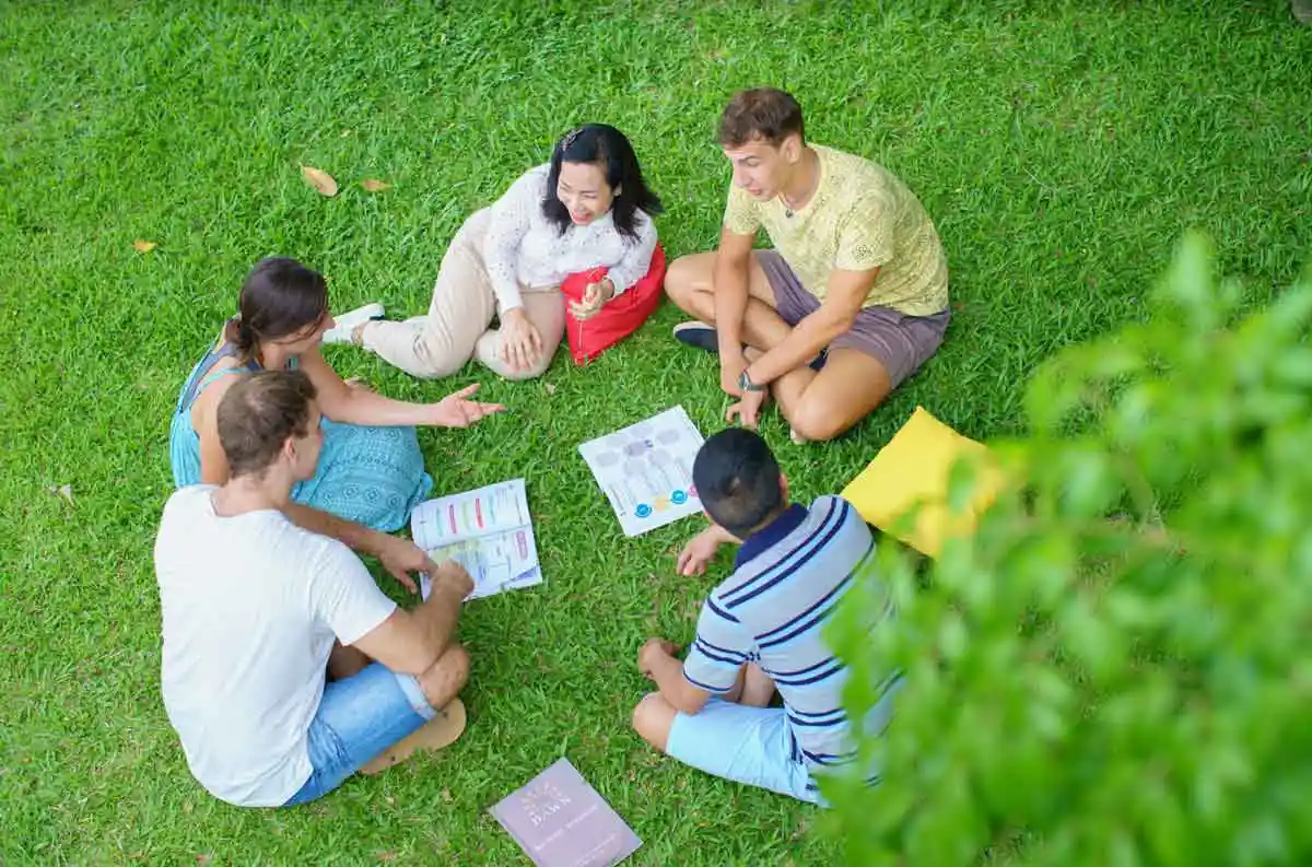 A group of five people sitting on the grass in a circle, engaging in a discussion with papers and materials spread out between them, enjoying an outdoor setting at The Dawn.