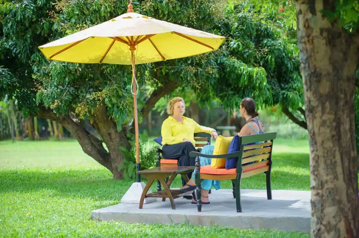 Two people sitting on a bench under a yellow umbrella, talking in a lush garden setting with trees in the background at The Dawn.