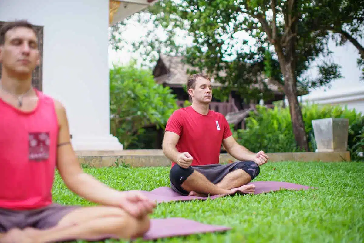 2 men meditating in the Easy Pose on a yoga mat in a garden at The Dawn.