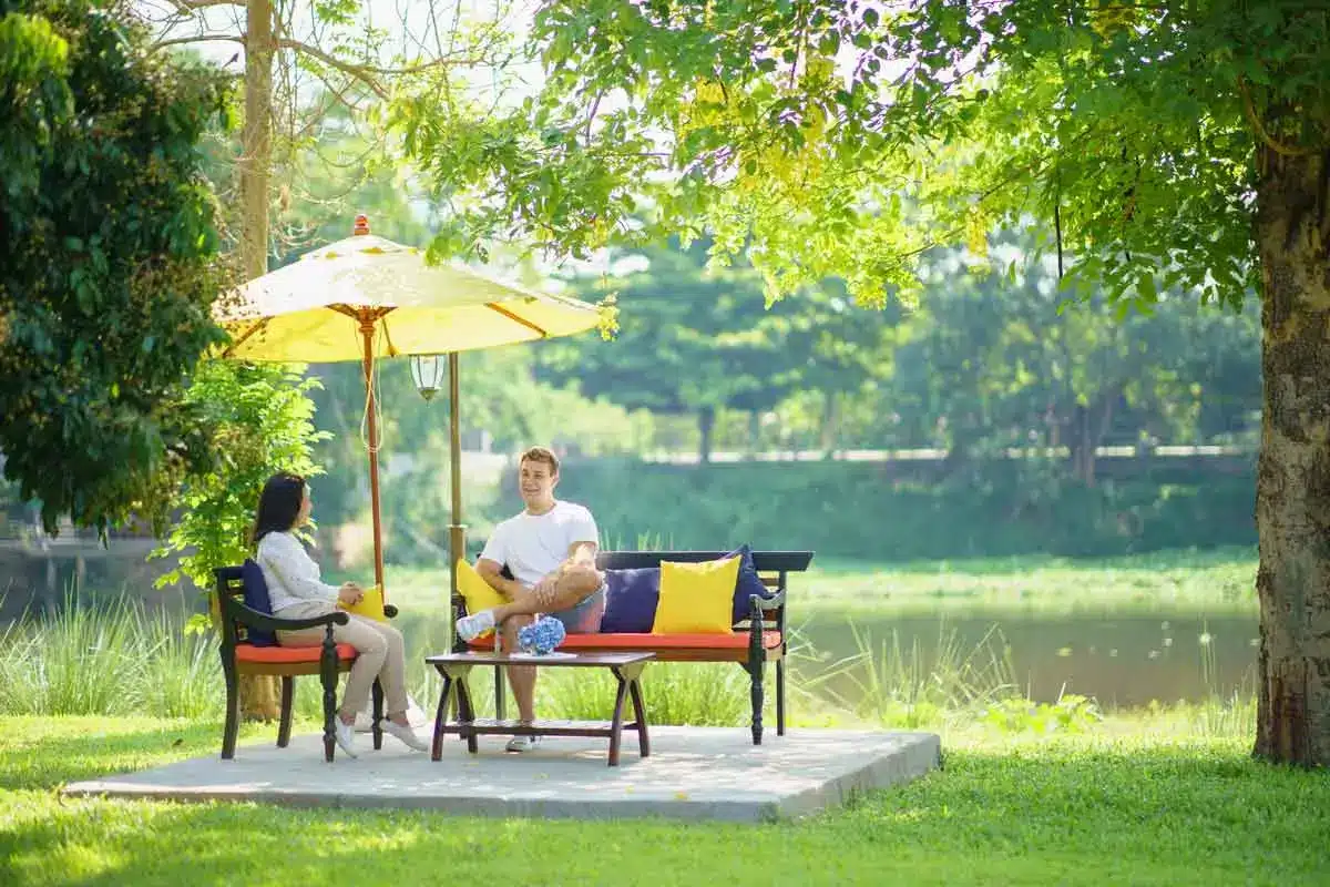 Two people talking, seated on benches under a yellow umbrella by the river at The Dawn.