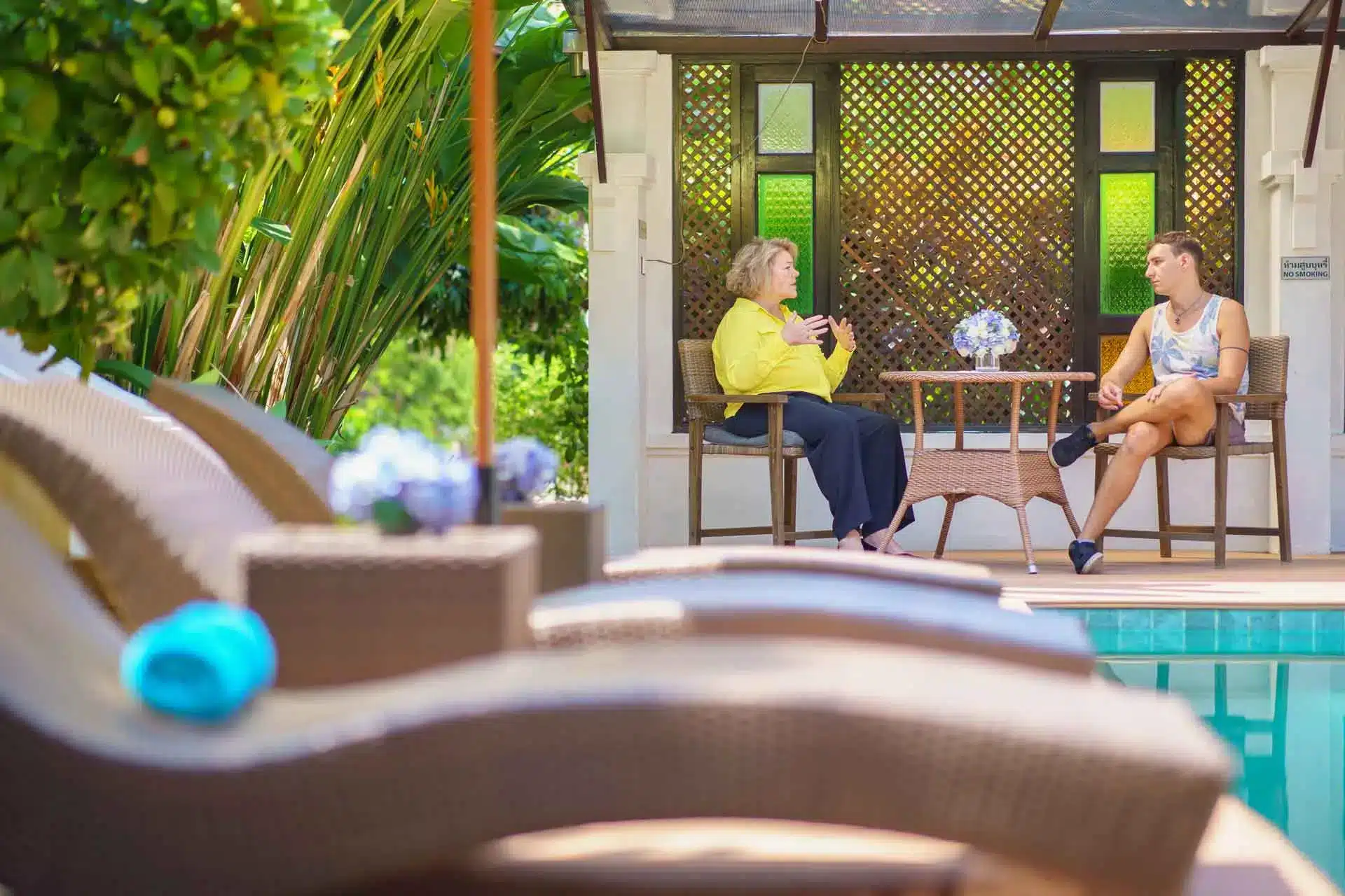 Two people talking by a poolside seating area, surrounded by lush greenery and a wooden lattice screen in the background.