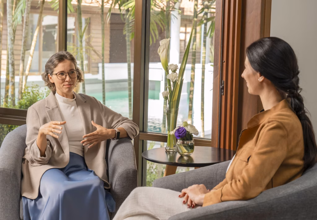 A female therapist engages in a supportive conversation with a female client during a private session in a calm, sunlit room.