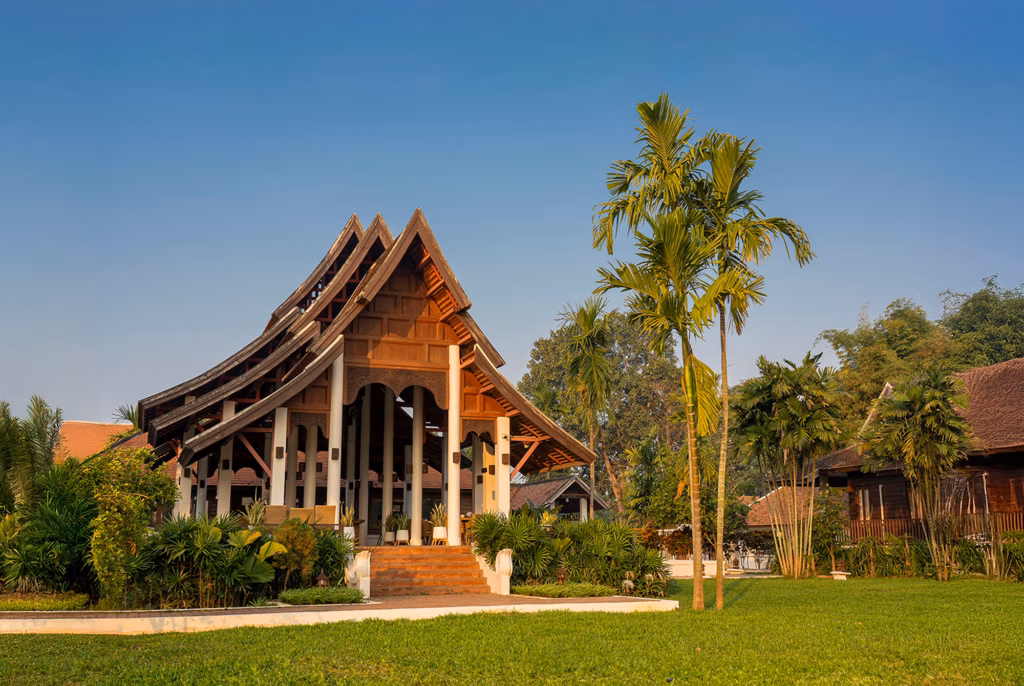 A traditional Northern Thai-style wooden pavilion with a steep, tiered roof, surrounded by palm trees and a lush green lawn under a clear blue sky.