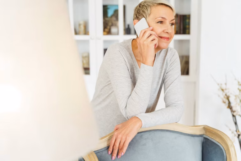 A woman with short blonde hair, wearing a light grey long-sleeved top, smiles while talking on a white smartphone. She leans on the back of a pale blue upholstered armchair in a bright living room, with a white bookcase and a lampshade blurred in the foreground.