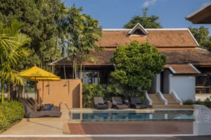 A rectangular outdoor swimming pool bordered by stone paving and wicker sun loungers with navy towels, set in front of a traditional Thai Lanna-style building with a steep terracotta tile roof and white trim. A yellow patio umbrella shades loungers on the left, tall palms and a large rounded shade tree fill the garden, and a wide staircase with curved white balustrades leads up to the building on the right, all under a clear blue sky.
