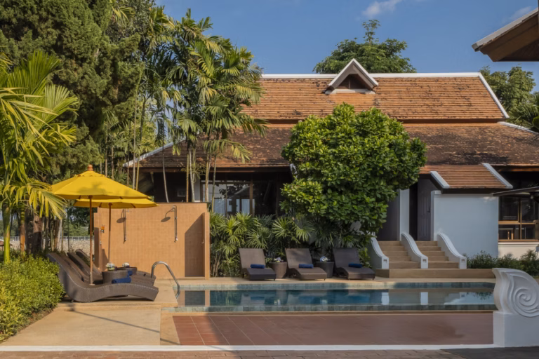 A rectangular outdoor swimming pool bordered by stone paving and wicker sun loungers with navy towels, set in front of a traditional Thai Lanna-style building with a steep terracotta tile roof and white trim. A yellow patio umbrella shades loungers on the left, tall palms and a large rounded shade tree fill the garden, and a wide staircase with curved white balustrades leads up to the building on the right, all under a clear blue sky.
