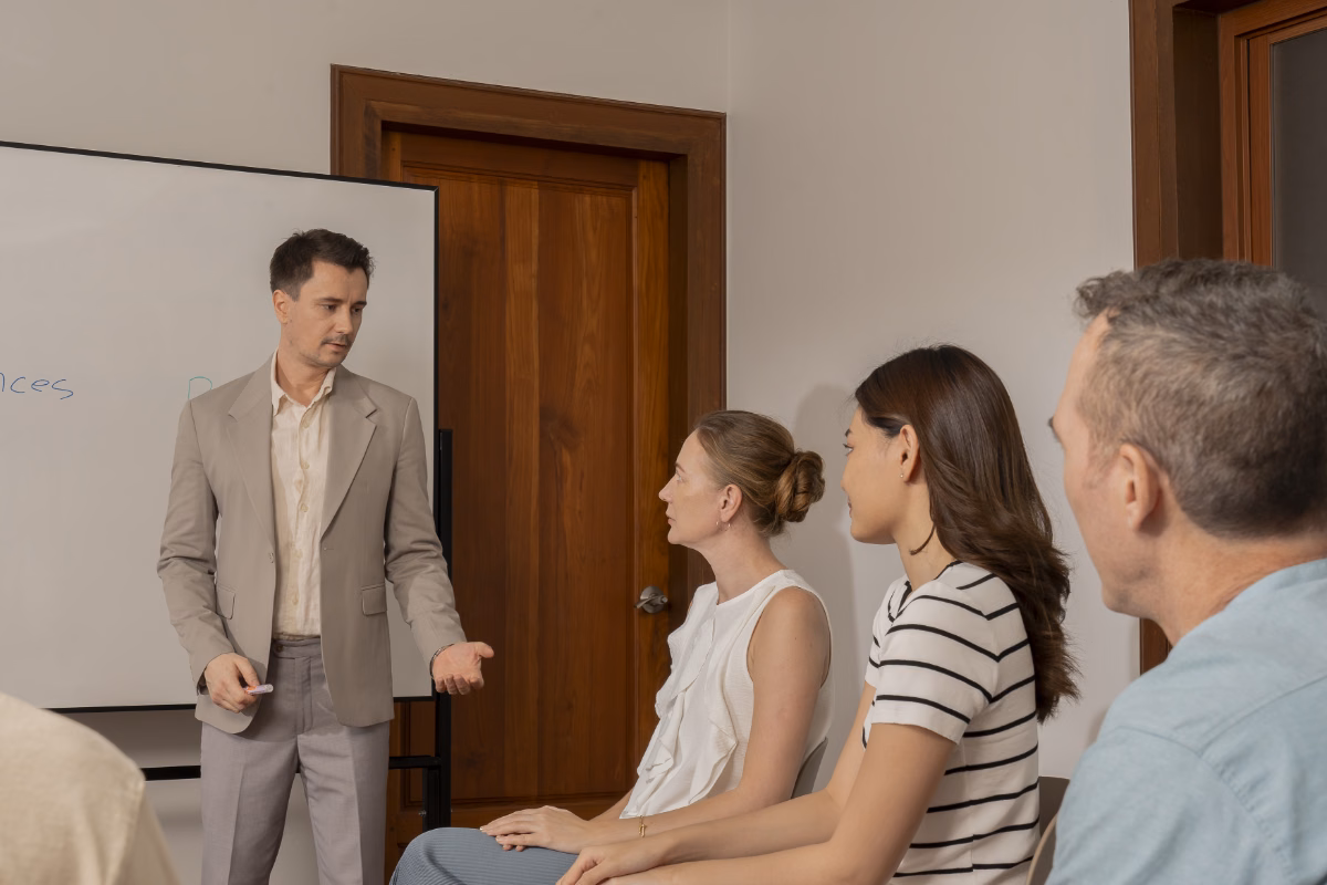 A man in a light grey suit stands beside a whiteboard in a conference room, gesturing with an open hand as he speaks to a seated group of colleagues, including two women and a man in a light blue shirt who are turned toward him and listening.