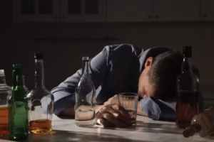 A man in a blue denim shirt slumped face-down on a kitchen table in a dimly lit room, one hand resting beside a half-full drinking glass, surrounded by numerous empty and partially full liquor and beer bottles.