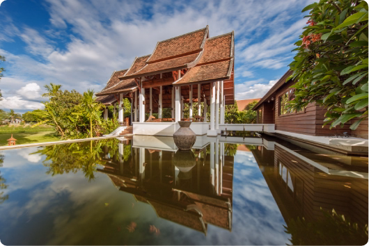Thai-style teak pavilion reflected in the pool under a blue sky at The Dawn Rehab Thailand