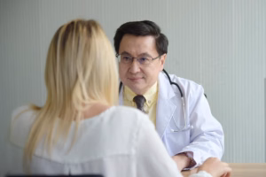 A male doctor wearing glasses, a white lab coat, and a stethoscope listens attentively across a desk to a blonde female patient seen from behind.